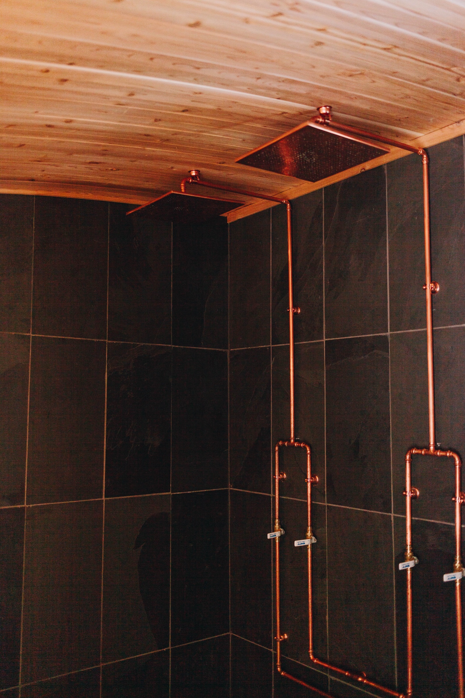 Copper rain shower fixtures with exposed piping in dark-tile two-person shower room